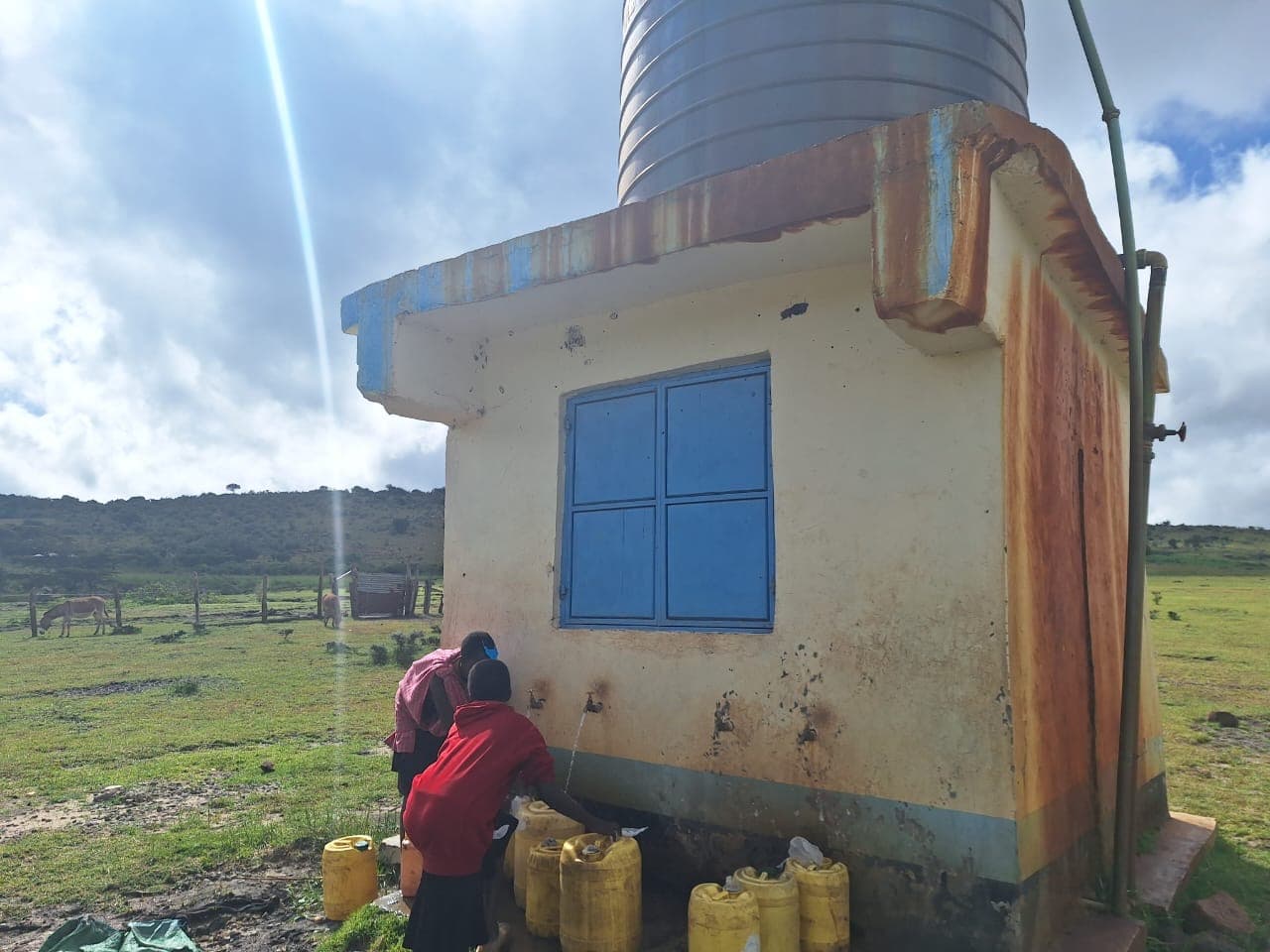 Dos niñas llenan garrafas amarillas en el punto de agua actual, junto a un depósito oxidado